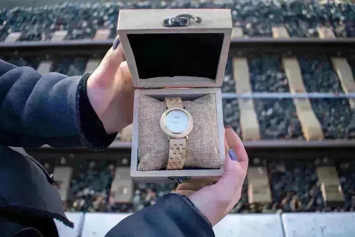Hands holding an open wooden watch box featuring a light wood timepiece on a textured beige cushion.