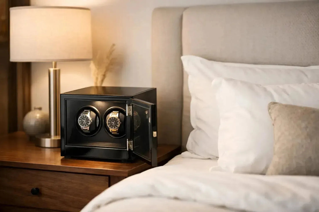 Black wooden double watch winder on a bedside table next to a lamp and a cozy bedroom headboard.