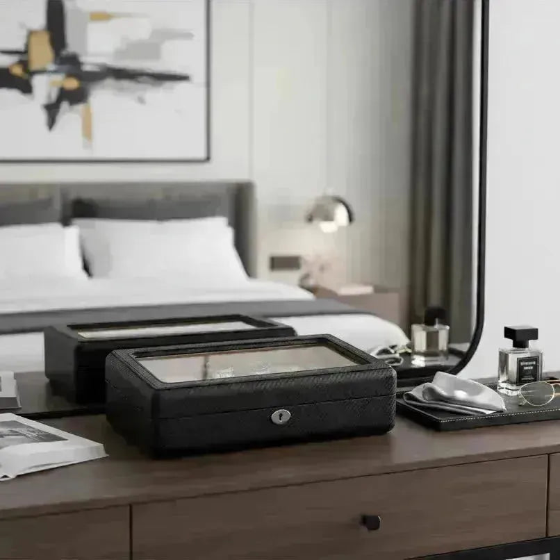 Black leather watch box with a glass top on a wooden dresser, reflecting a modern bedroom.