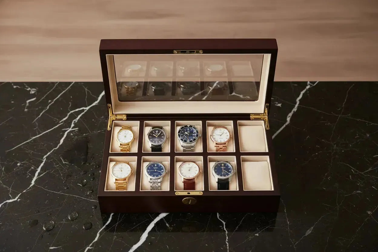 Dark wooden watch display case with a glass lid, holding eight luxury wristwatches on cream velvet cushions.