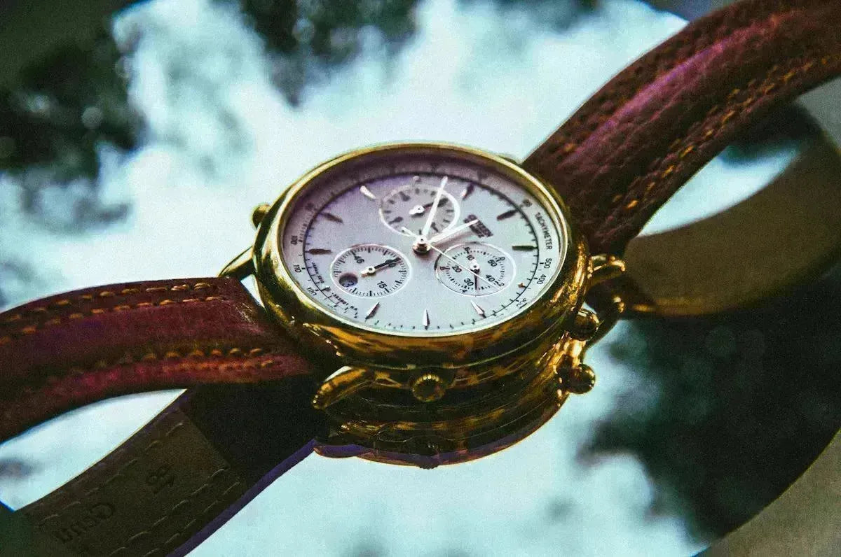 Elegant gold chronograph watch with a brown leather strap resting on a reflective surface.