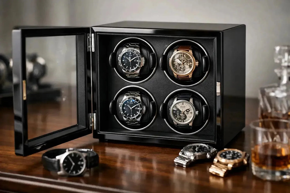 Black wooden watch winder box with glass door holding four luxury timepieces on a polished desk with whiskey glasses.