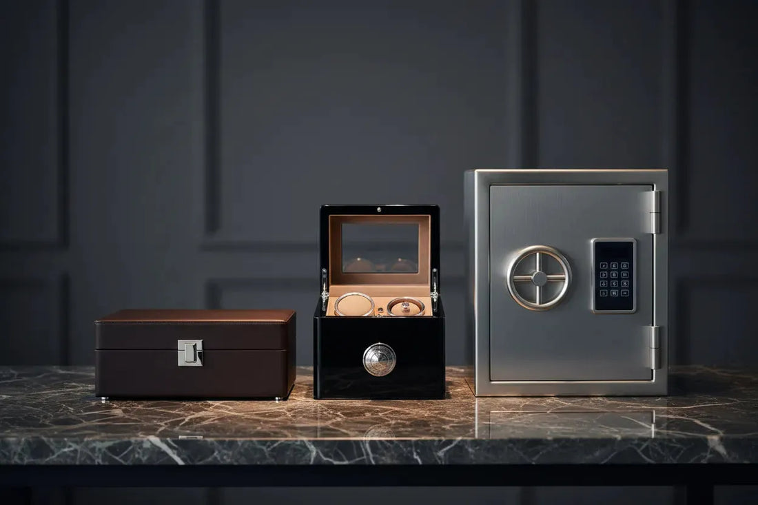 Brown watch box, open black watch winder, and a silver safe on a marble surface.