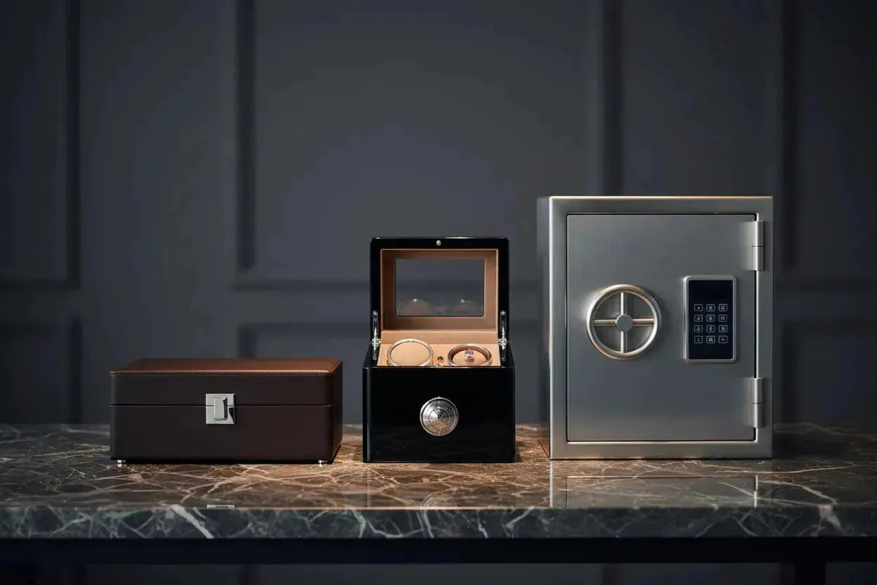 Brown leather watch box, black automatic watch winder, and metallic digital safe arranged on a marble surface.
