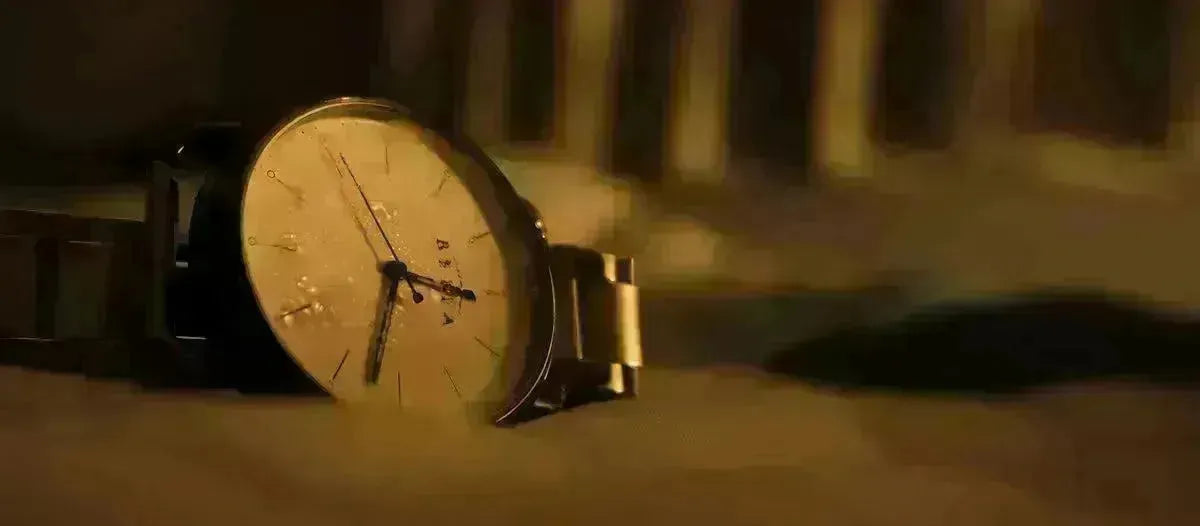 Luxury analog wristwatch with a metallic band resting on a soft, warm-toned surface in dim lighting.