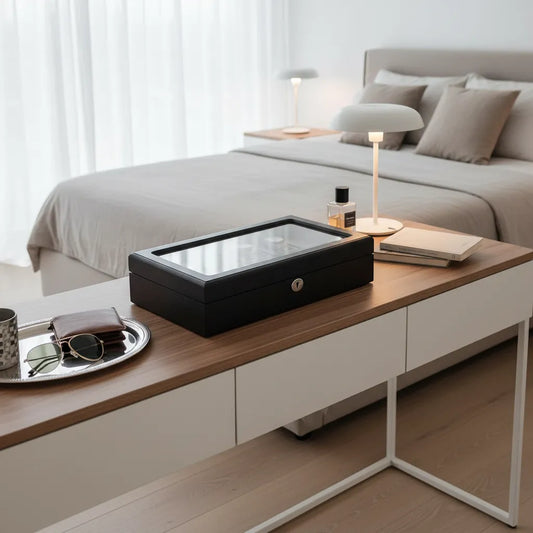 A modern, minimal bedroom with a beige bed, soft pillows, and a wooden desk displaying the Tempus Luxury Watch Winders 12 Watch Box Premium Black Oak Veneer, plus books, a lamp, sunglasses, wallet, and cup by a large window with sheer curtains.