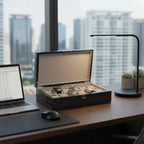 Black carbon fibre 12-watch box with curved lid open, showcasing three watches on a desk with a laptop.