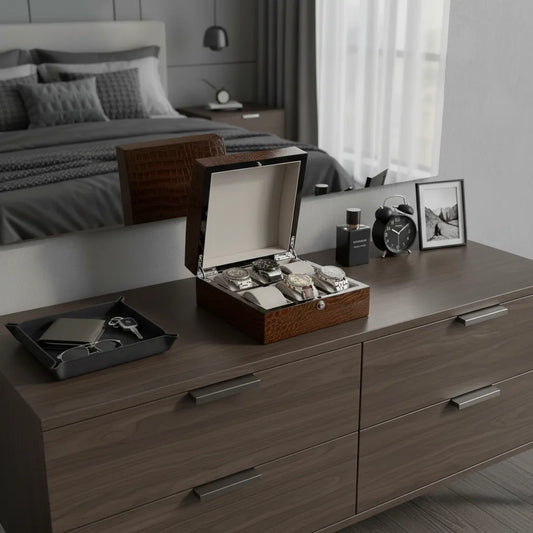 A modern bedroom dresser displays the Tempus Luxury Watch Winders 6 Watch Box in Brown High Gloss Lacquered Crocodile Print Finish, alongside a jewelry and phone tray, framed photos, alarm clock, and perfume bottles. A spotless bed and window are in view.