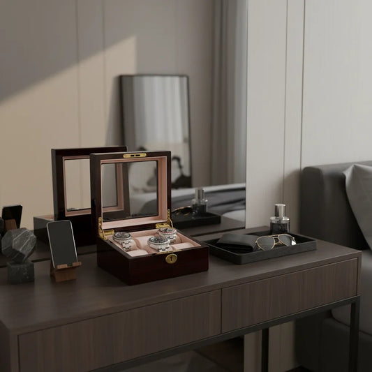 A Tempus 6 Watch Box in Premium Cherry Wood Piano Gloss Finish with Luxury Lining, displaying two watches, sits on a modern desk beside a phone stand, dark tray with sunglasses, bottle, decor, mirror, and sofa in the background.