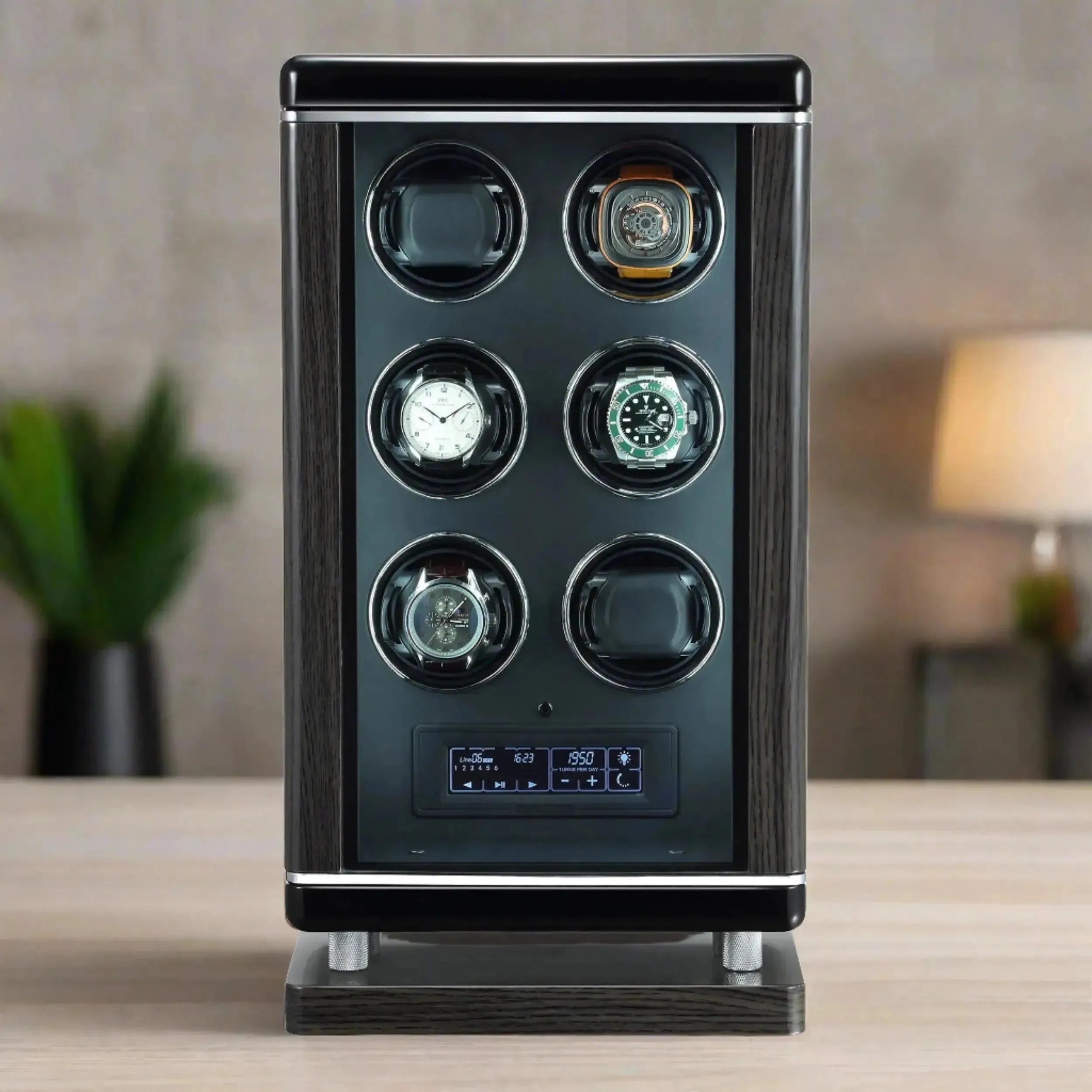 The Tempus Luxury 6 Watch Winder for Automatic Watches with BioMetric Technology holds three watches, features a digital display panel at the bottom, and sits on a wooden surface with a blurred plant and wall in the background.