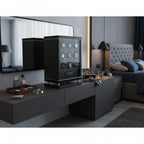 A modern bedroom features the 9 Watch Winder for Automatic Watches with BioMetric Technology by Tempus Luxury Watch Winders on a dark wood vanity, alongside sunglasses, a watch, and drinks, with a tufted headboard and lit wall sconce in the background.