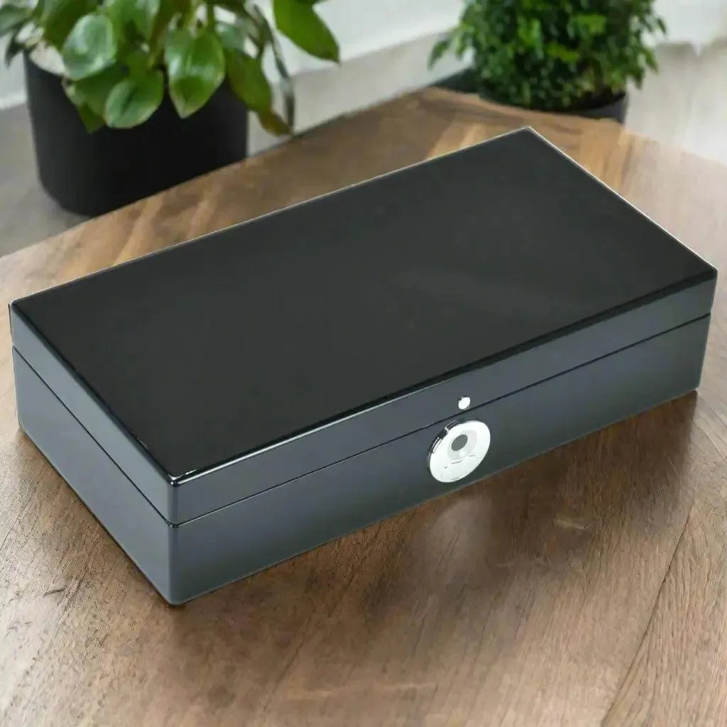 The Tempus 10 Watch Box in Black Piano Finish with a Bio Metric Lock by Tempus Luxury Watch Winders rests on a wooden table, its sleek rectangular design surrounded by green potted plants in the background.