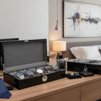 A modern bedroom showcases the Tempus Luxury Watch Winders' Tempus 10 Watch Box in Black Piano Finish with Bio Metric Lock, displaying watches, cologne, a wallet, cufflinks, a blue tie, and another box atop a wooden dresser amid stylish decor.