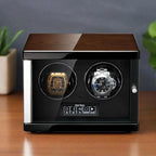 The Tempus 2 Watch Winder Automatic by Tempus Luxury Watch Winders features a wooden top, black sides, and a glass front displaying two luxury watches; it rests on a wooden surface with a blurred plant in the background.