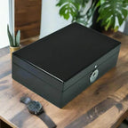 The Tempus 8 Watch Box in Black Piano Finish with Bio Metric Lock by Tempus Luxury Watch Winders rests on a wooden table, accompanied by a luxury watch and a small potted cactus, with lush green plants in the background.