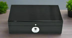 The Tempus 8 Watch Box in Black Piano Finish with Bio Metric Lock by Tempus Luxury Watch Winders sits closed on a wooden surface, flanked by small green potted plants—perfect for luxury watch storage.