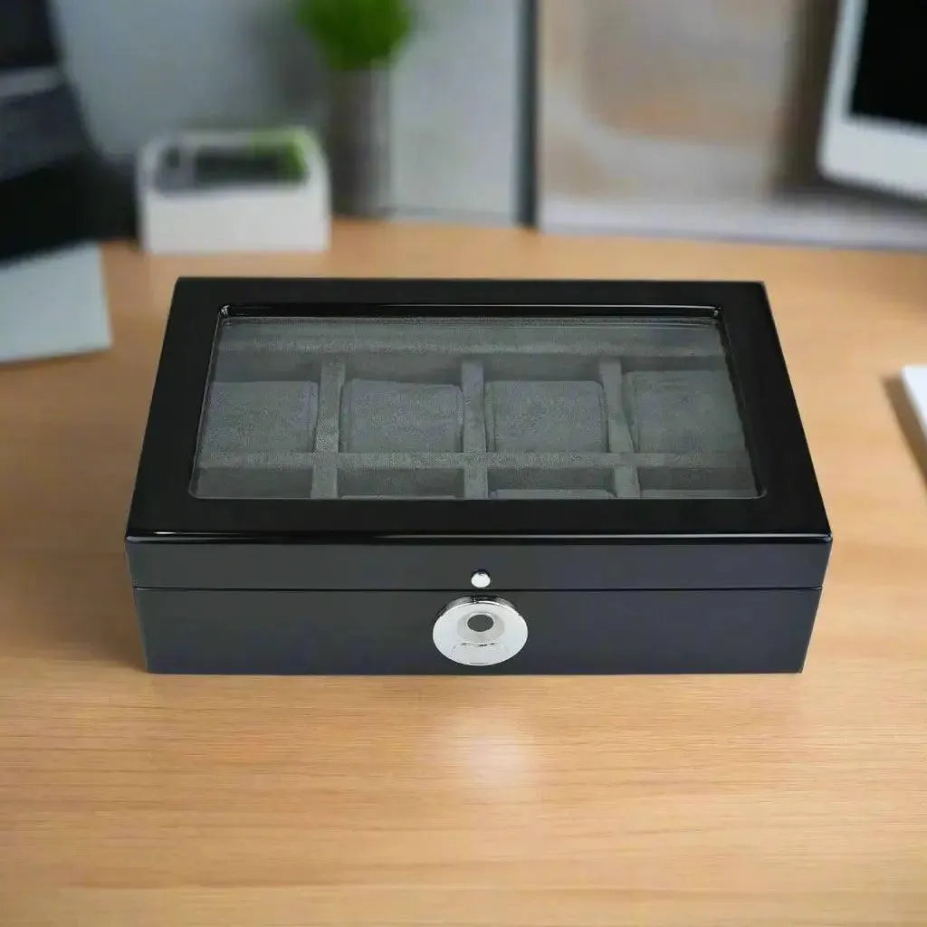 The Tempus 8 Watch Box in Black Piano Finish by Tempus Luxury Watch Winders, featuring a glass lid, interior compartments, and a silver biometric lock on the front, sits on a wooden desk against a background of blurred office items.