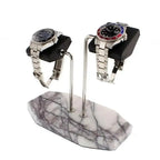 Two stainless steel watches rest on black leather cushions atop the Tempus Double White Marble Watch Stand with dark violet veins by Tempus Luxury Watch Winders, elegantly supported on a white marble base.