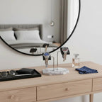 A wooden dresser displays sunglasses, a wallet, cologne, polka-dot handkerchiefs, and two wristwatches on the Tempus Double White Marble Watch Stand Dark Violet Veins with Black Leather by Tempus Luxury Watch Winders. A round mirror reflects a bed and curtains.