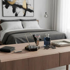 A modern bedroom features a neatly organized wooden desk displaying a luxury gift: the Tempus Watch Stand in Black Marble Brown Veining with Black Leather by Tempus Luxury Watch Winders, along with glasses, wallet, blue perfume bottle, and tray. Gray linens and abstract art are in the background.
