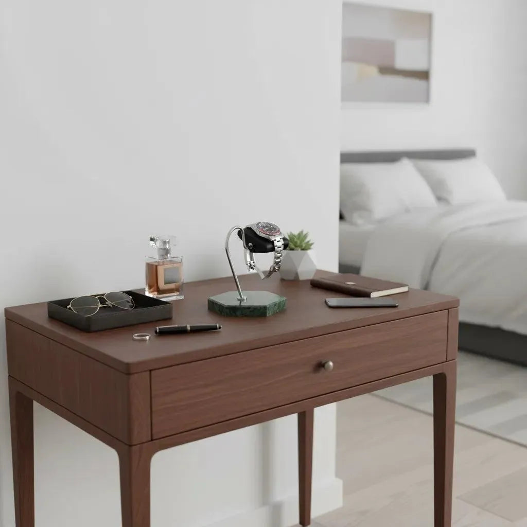 A wooden bedside table with a drawer displays sunglasses, a perfume bottle, and a wristwatch on the Tempus Watch Stand in Green Marble Hexagon Base with Black Leather by Tempus Luxury Watch Winders. A pen, two closed notebooks, and wall art are also visible.
