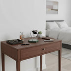 A wooden bedside table with a drawer displays sunglasses, a perfume bottle, and a wristwatch on the Tempus Watch Stand in Green Marble Hexagon Base with Black Leather by Tempus Luxury Watch Winders. A pen, two closed notebooks, and wall art are also visible.