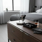 A modern bedroom features the Tempus White Marble Watch Stand (Dark Violet Veins, Black Leather) by Tempus Luxury Watch Winders on a wooden dresser, displaying a watch beside a tray with keys and a ring, cologne bottle, and glasses. A tidy bed sits by the window.
